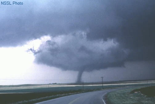 A dark, rounded lowering hangs beneath a storm base over open farmland, with a narrow cone-shaped tornado extending from it to the ground and a lighter slot cutting into the cloud base beside it